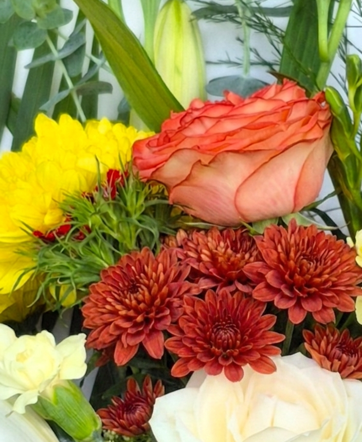 A professional photograph of a Florist Choice bouquet in a rich sienna palette, featuring brick-red and yellow chrysanthemums, peach and white roses, white carnations, and lily buds, nestled in eucalyptus and magnolia foliage.